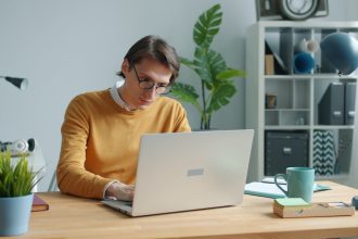Man in yellow sweater working on laptop at desk; downtime