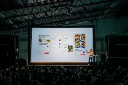 person discussing while standing in front of a large screen in front of people inside dim-lighted room; Pitch Your Services