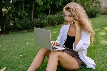Young woman enjoying remote work on a laptop while talking on a phone outdoors in a sunlit park.; productive during slow months