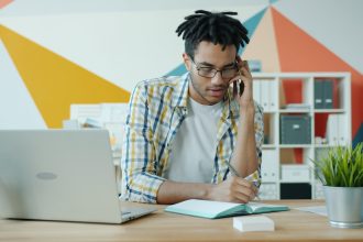 Man talking on phone at desk with laptop and notebook.; vetting business tools for freelancers