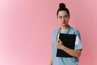 Confident female doctor in scrubs holding a clipboard against a pink background.; online nursing program