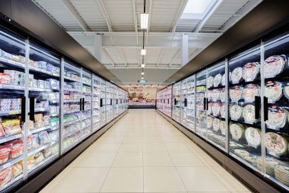 A long aisle in a brightly lit supermarket with refrigerators.; boring store aisles
