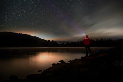 a person standing on a rocky shore with a body of water and stars in the sky; professional flashlights