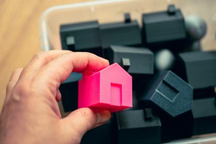 A person holding a pink house in front of a pile of black cubes; May housing starts