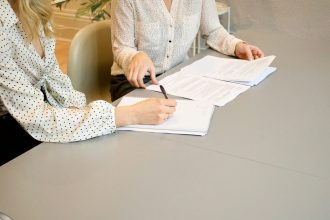 woman signing on white printer paper beside woman about to touch the documents; proforma invoice