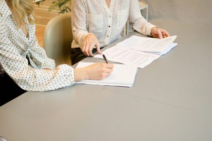 woman signing on white printer paper beside woman about to touch the documents; proforma invoice