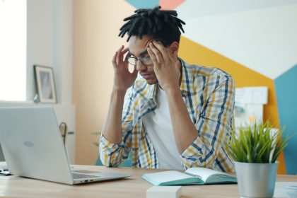 Man with dreadlocks holding head at desk with laptop; jobless claims