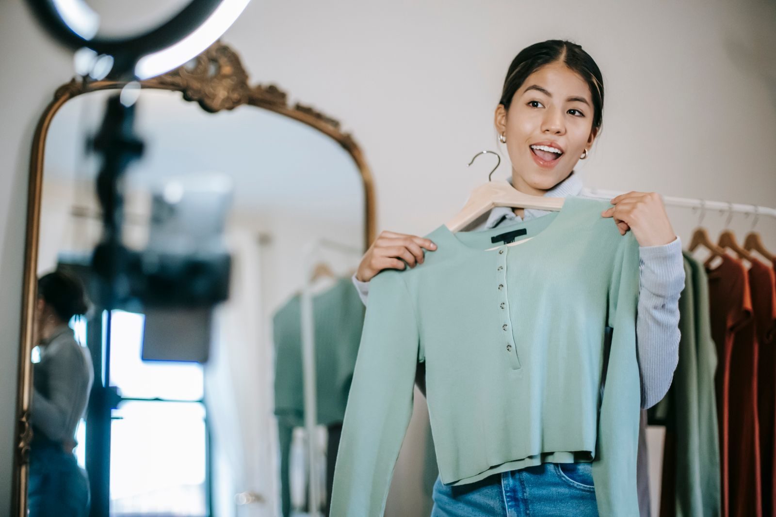 Smiling young ethnic female blogger standing in room near wardrobe and mirror while demonstrating blouse on hanger and filming vlog on mobile with ring lamp; creator campaigns