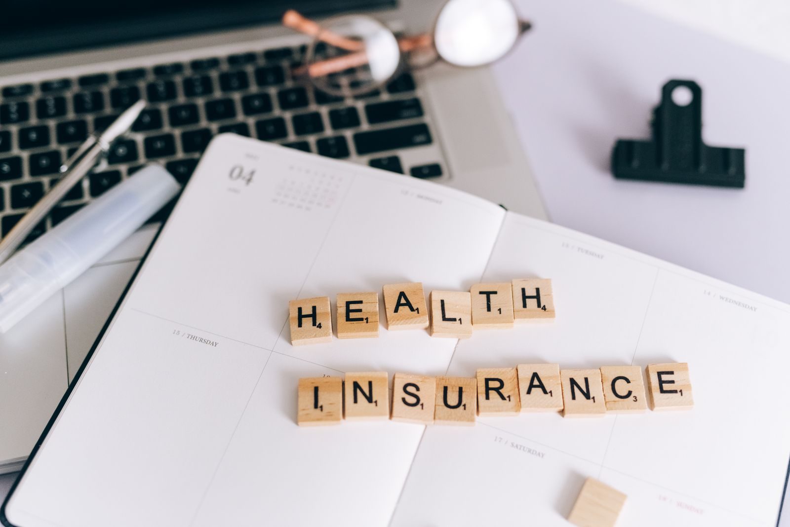 Close-up of Scrabble tiles spelling 'Health Insurance' on a planner with a laptop; small business health insurance premiums 2027