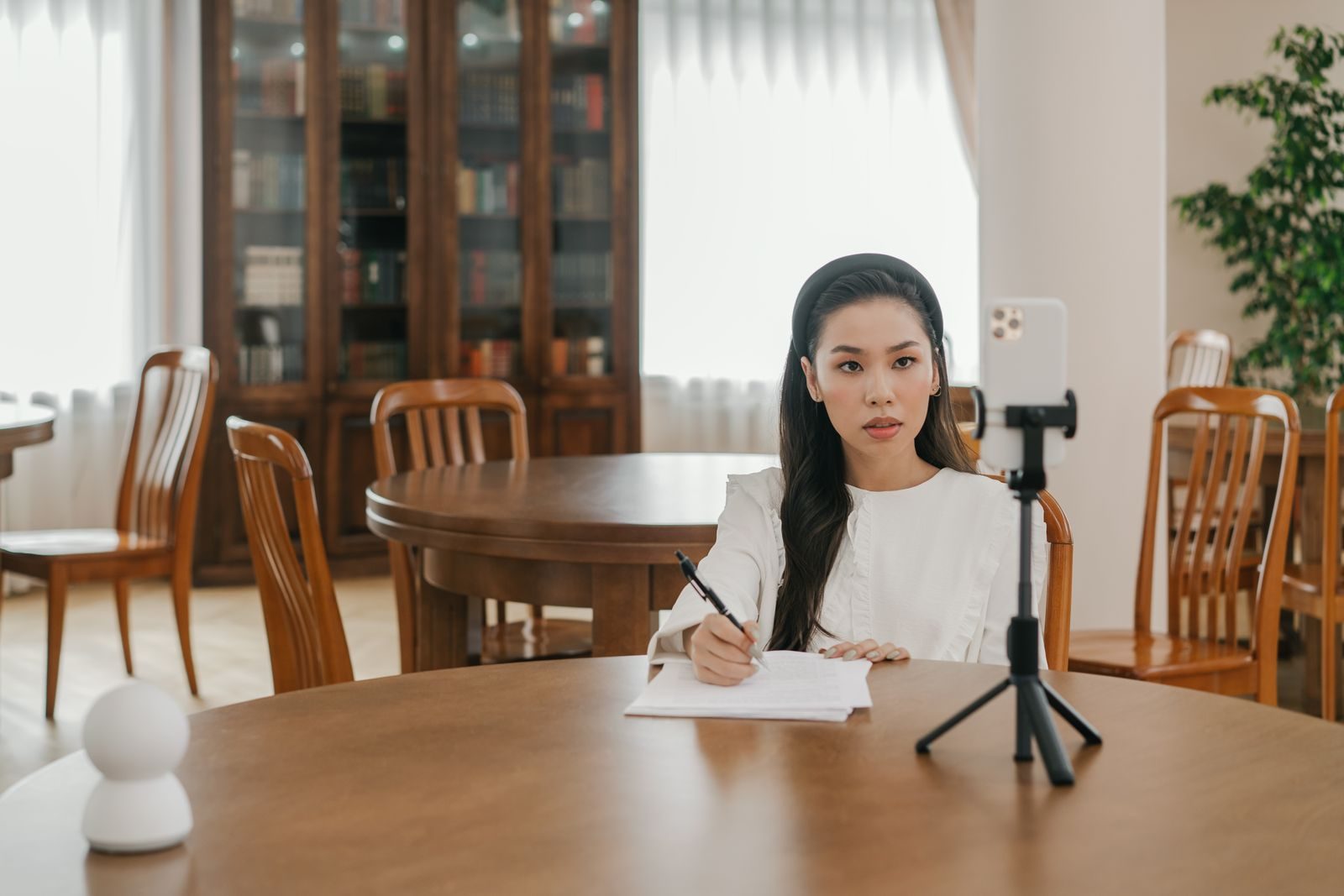 Young Asian woman vlogging in a cozy home office setup using a smartphone and tripod; AI creator outreach