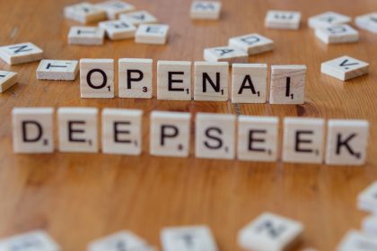 Close-up of wooden Scrabble tiles spelling OpenAI and DeepSeek on wooden table; OpenAI dots