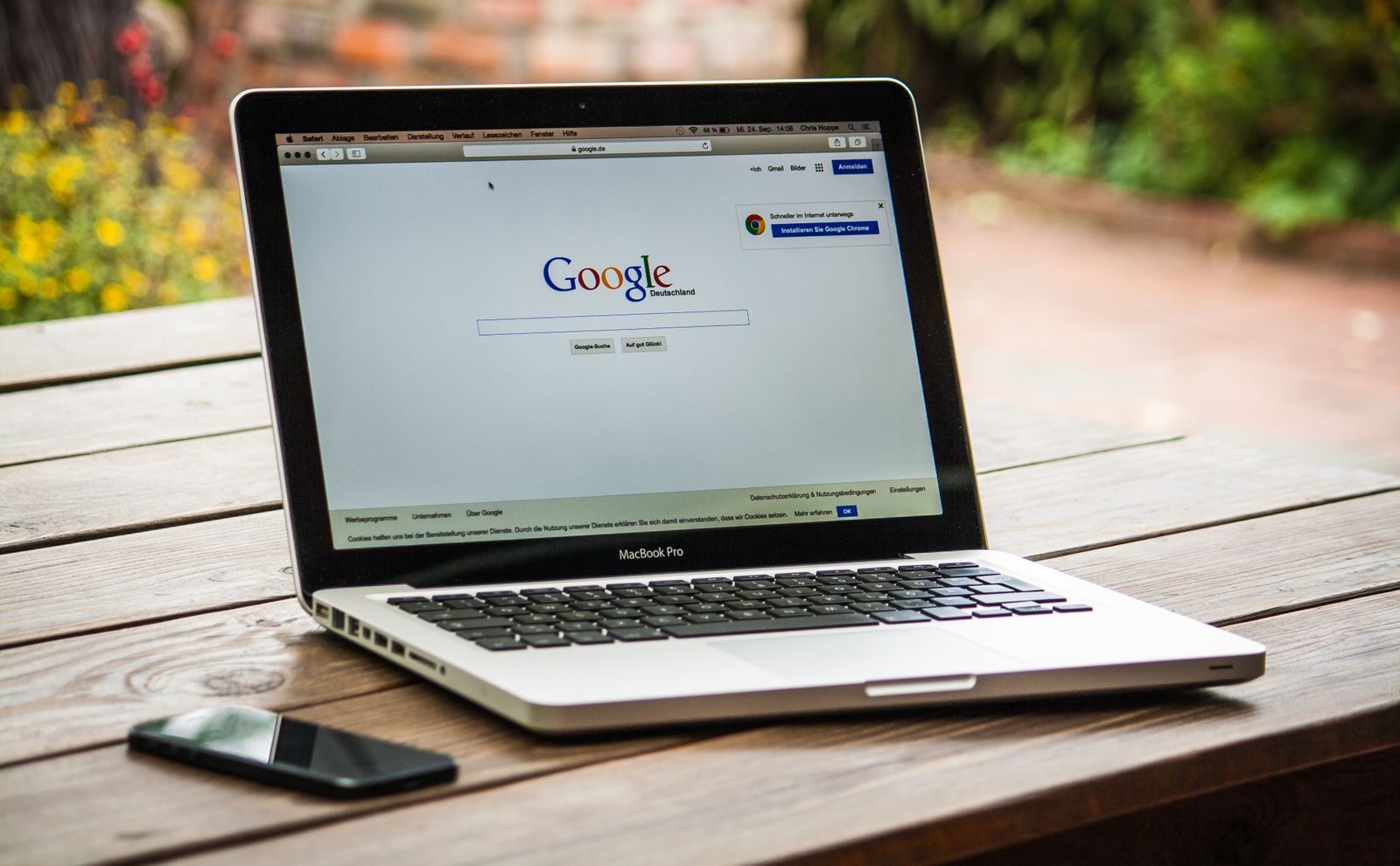 A MacBook Pro displaying Google Search on a wooden table outdoors, next to a smartphone; Google Deep Review