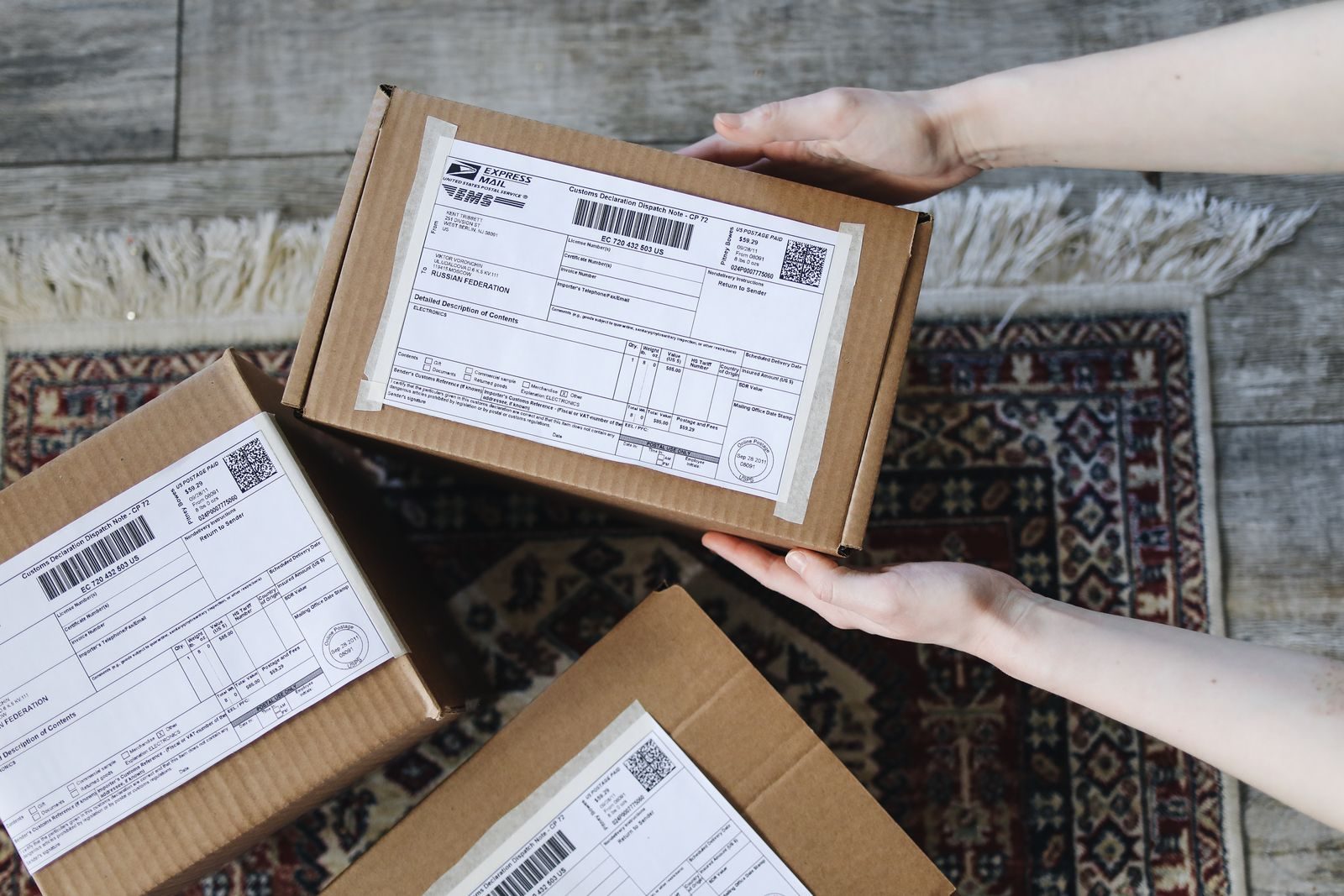Close-up of hands holding cardboard boxes with labels for delivery on a decorative carpet; Poshmark overweight shipping