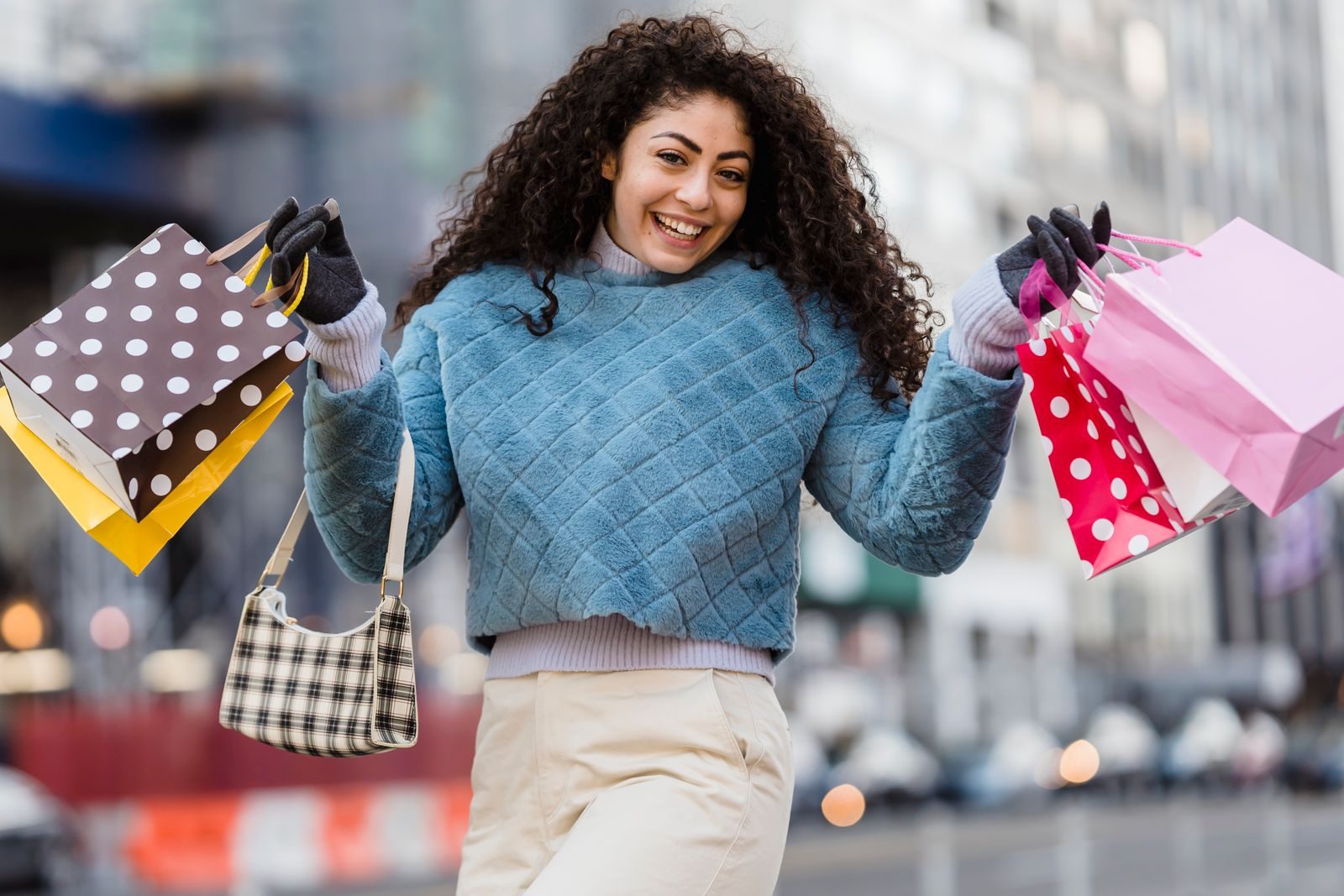 Positive young female in stylish outfit with colorful gift bags smiling and looking at camera on blurred background of street; consumer confidence