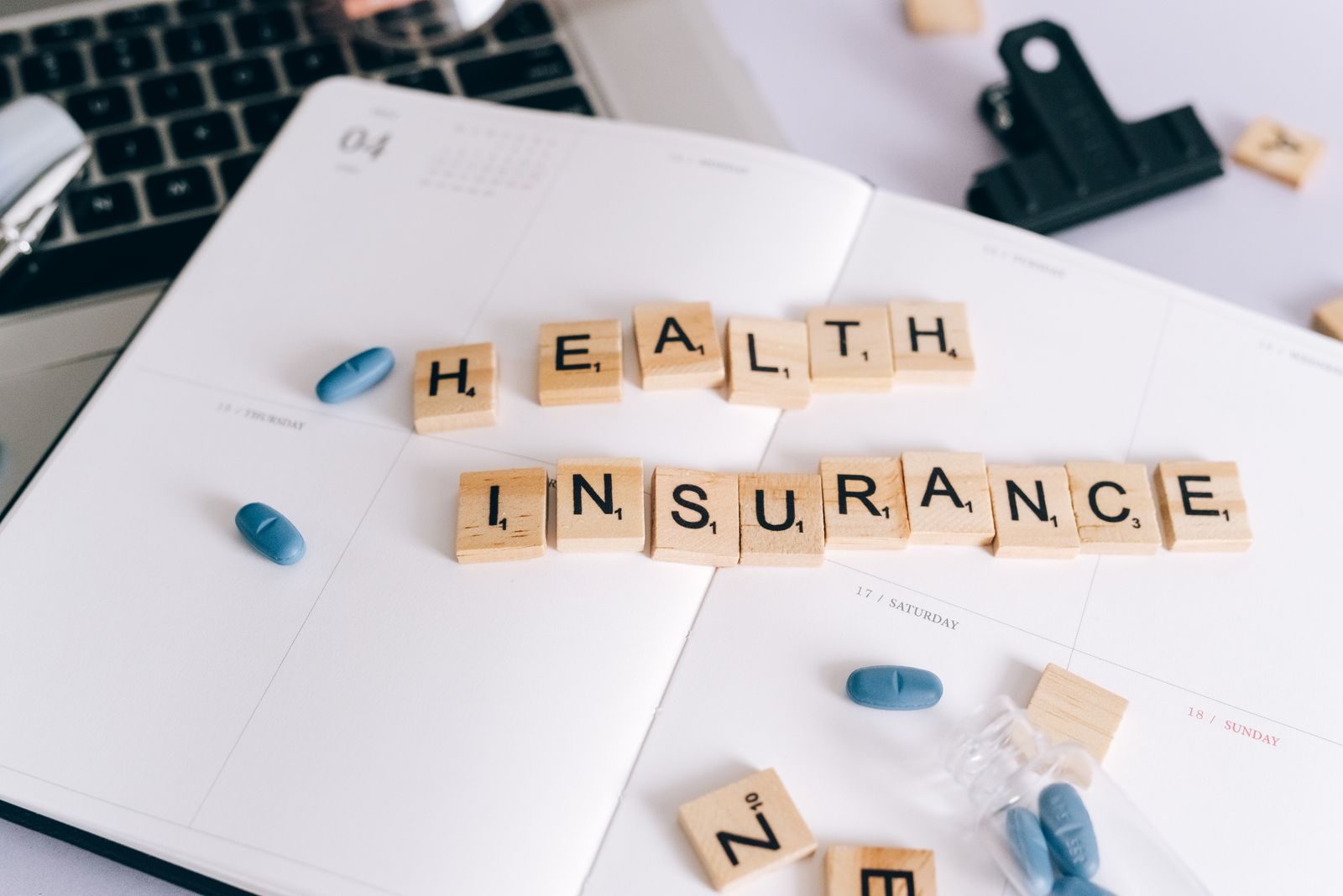 Scrabble tiles spelling 'Health Insurance' on planner with pills and laptop, symbolizing healthcare planning; NFIB health coverage