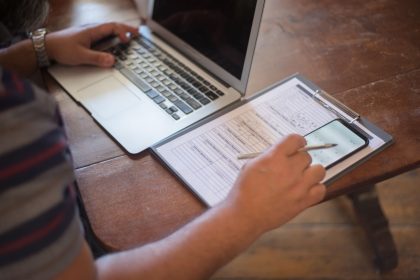 Close-up of a person using a laptop and reviewing documents with a smartphone on a wooden table; Small Business Check Up Survey