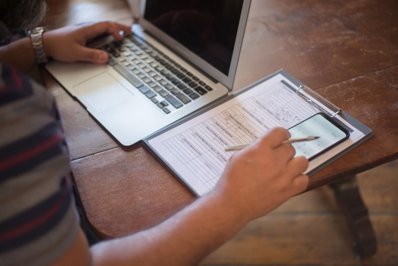 Close-up of a person using a laptop and reviewing documents with a smartphone on a wooden table; Small Business Check Up Survey